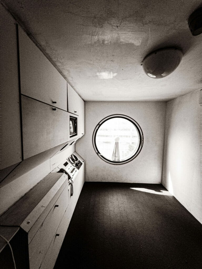 An interior view of an empty, unfurnished capsule. There is a circular window at the opposite end, with built-in shelving and appliances on the lefthand side.